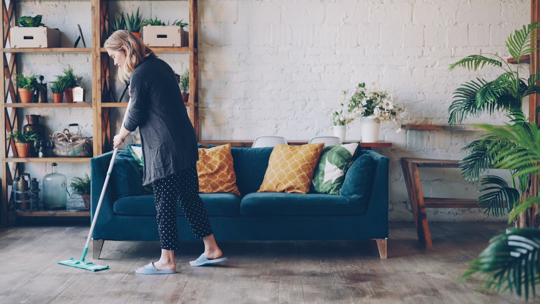 Attractive young woman is cleaning living room mopping the floor doing housework. Beautiful loft style apartment with modern furniture and green plants is visible.